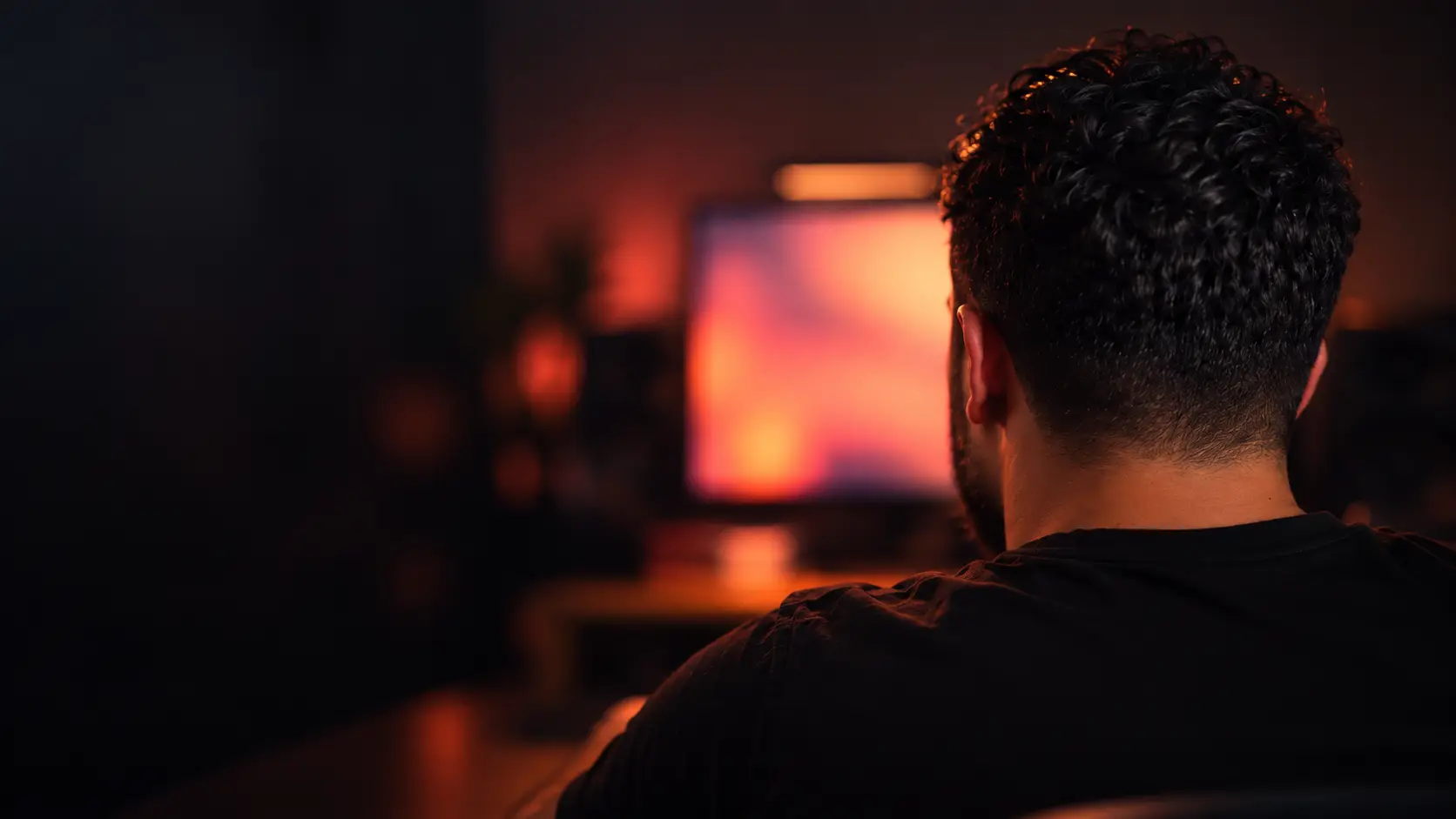 Pessoa de costas diante de um monitor aceso, desfocado, numa sala escura com luz quente em coral e rosa.